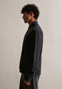 Čierna dlhá polo košeľa s plochým golierom a gombíkovou légou, s voľným strihom. Nosená s tmavými džínami.