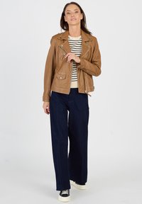 Braune Lederbikerjacke mit silberner Hardware, ausgestattet mit Reißverschlüssen und Taschen. Kombiniert mit marinenweiten Jeans und schwarzen Sneakern.