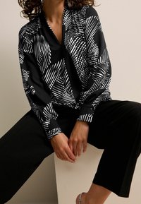 Sort bluse med et geometrisk hvidt mønster, lange ærmer med knapmanchetter, parret med sorte bukser og sølv sandaler.