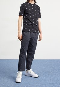 Schwarzes Baumwoll-T-Shirt mit wiederholt weißen und lila Nike-Logos, kombiniert mit grauen Hosen und weißen Nike-Sneakern mit schwarzen Akzenten.