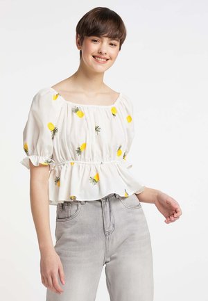 Giovane donna sorridente che indossa una camicetta bianca con stampe di ananas gialli e jeans a vita alta grigio chiaro su sfondo bianco.