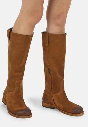 Braune kniehohe Wildlederstiefel mit seitlichem Reißverschluss, getragen an nackten Beinen, mit abgerundeten Zehen und niedrigen flachen Absätzen.
