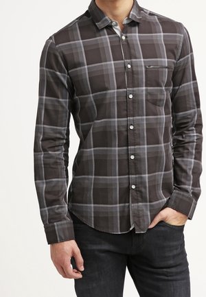 Mężczyzna w ciemnoszarej, zapinanej na guziki kraciastej koszuli z długim rękawem i kieszenią na piersi, w czarnych spodniach, z ręką w kieszeni.