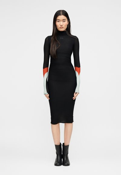 Γυναίκα με μακριά σκουρόχρωμα μαλλιά φοράει μαύρο εφαρμοστό midi φόρεμα με ψηλό λαιμό και αντίθετες μακριές μανσέτες σε κόκκινο και ανοιχτό μπλε, συνδυασμένο με μαύρα μποτάκια αστραγάλου.