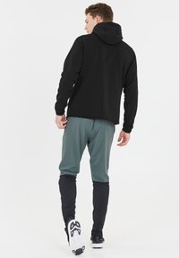 Sort hættetrøje med et enkel design, parret med lysegrå bukser med sorte detaljer. Model vender væk, hvilket viser tøjsets pasform.