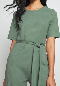Geribbelde groene jumpsuit met korte mouwen, een ronde halslijn en een strikceintuur in de taille. Zachte stof met een gestructureerd vlak.