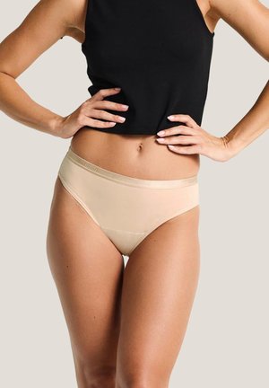 Frau trägt beige nahtlose Unterwäsche und ein schwarzes ärmelloses Oberteil, die Hände an der Taille ruhend vor einem schlichten Hintergrund.