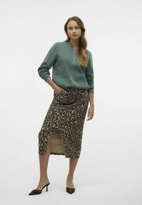 Sweatshirt zippé vert, jupe midi imprimé léopard avec une fente sur le côté, et escarpins marron à bout pointu avec des accents décoratifs. Textures lisses.