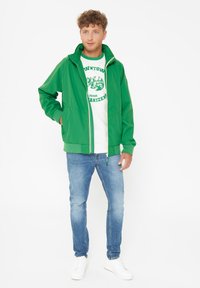Green zip-up jacket with a hood, featuring elastic cuffs and bottom hem. Worn over a white t-shirt with green graphics, paired with light blue jeans.