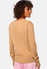 Beige gestrickter Pullover mit langen Ärmeln, rundem Ausschnitt und geripptem Saum. Glatte Textur und lockere Passform, kombiniert mit rosa Hosen.