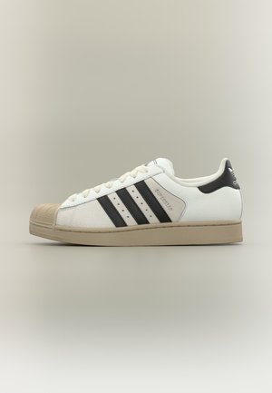 Baskets blanches Adidas Superstar avec bandes noires, semelle beige et bout texturé, présentées de profil sur un fond neutre.