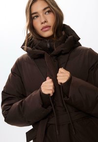 Smeđi puffer jakna s visokim ovratnikom, zatvaranjem na zip i pojasom na vezanje. Ima velike, mekane manžete i glatku, sjajnu teksturu.