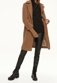 Brauner, strukturierten Mantel mit eingeschnittenem Kragen, schwarzer Kapuzenpullover mit Nieten und glänzenden schwarzen Kunstlederhosen, kombiniert mit schwarzen Schnürstiefeln.