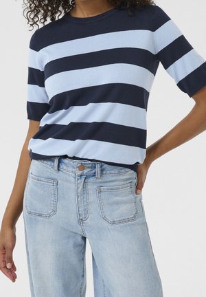 Nainen pukeutunut tummansiniseen ja vaaleansiniseen raidalliseen lyhythihaiseen puseroon, yhdistettynä vaaleanpesuihin korkeavyötäröisiin farkkuihin, joissa on etutaskut.