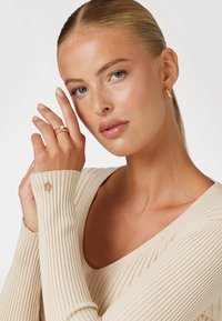 Beige ribbed sweater with long sleeves; model wears layered gold rings and hoop earrings. Smooth skin, natural makeup, neutral background.