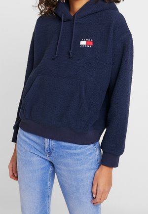 Felpa con cappuccio blu navy in pile, dotata di tasca a marsupio, orlo ribbed e coulisse regolabile. Presenta una piccola etichetta con logo in rosso, bianco e blu.