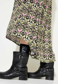 Zwarte leren enkellaarzen met een vierkante neus, gespen en studs, gecombineerd met een gemusterde groen-pink rok en contrasterende zwarte sokken.