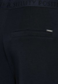 Granatowe spodnie dresowe mają teksturowany pas z napisem "POZYTEWNY" oraz małą metalową metkę na tylnej kieszeni.