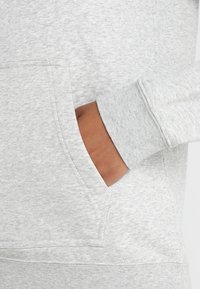 Šedá mikina s kapsou vpředu a žebrovanými manžetami, vyrobená z měkké, strukturované látky. Viditelné švy podél švů.