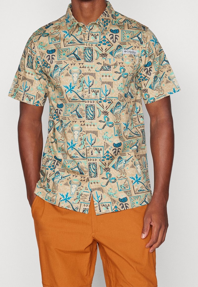 Homme portant une chemise beige à manches courtes avec des motifs naturels du sud-ouest en bleu et noir et un pantalon orange.