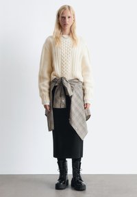 Flauschiger, cremefarbener Kabelstrickpullover, graues Karoshemd um die Taille gebunden, schwarzer Rock und schwarze Schnürstiefel mit dicken Sohlen. Minimalistischer Hintergrund.