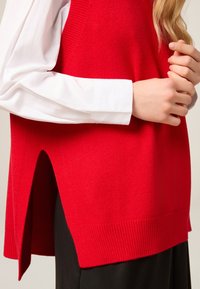 Roter Strickweste mit geripptem Saum, seitlichen Schlitzen und darüber getragen auf einem weißen Langarmshirt. Glatte Textur, lässiges Design.