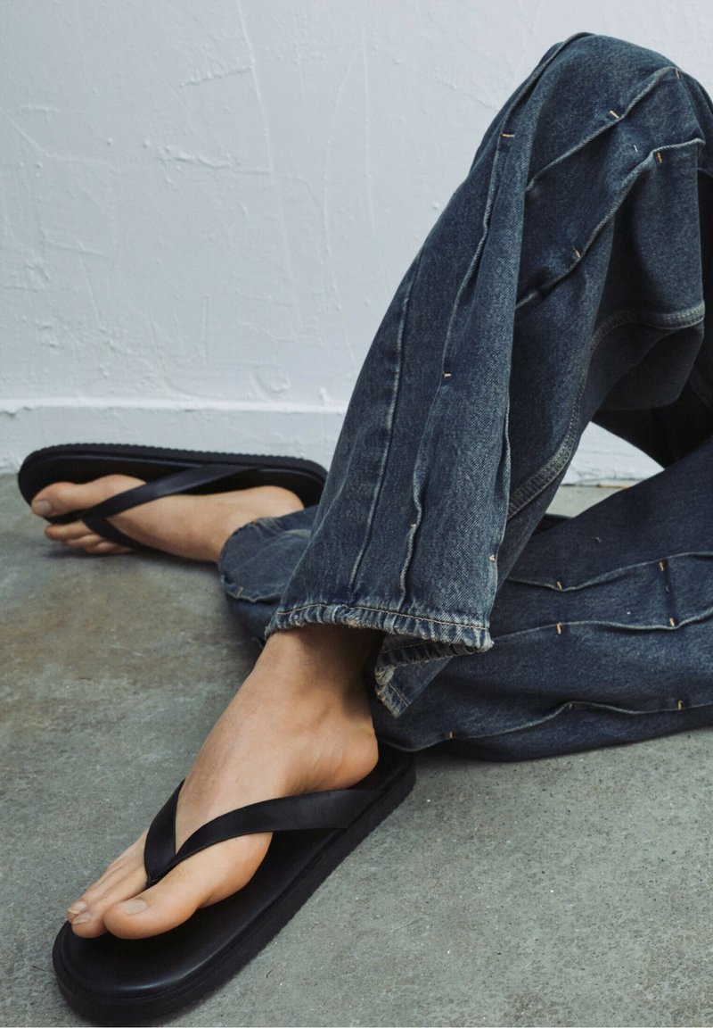 Infradito neri con una finitura opaca e liscia e larghe cinghie, abbinati a jeans di denim scuro, larghi e sfrangiati. Il terreno è di un grigio texturizzato.