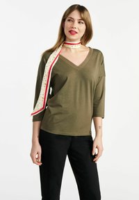 Oliwkowy top z dekoltem w serek i rękawami 3/4, ozdobiony kwiecistym szalem zawiązanym na szyi w czerwono-białe akcenty. Czarne spodnie.