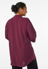 Burgundy long-sleeve shirt with a collar, textured fabric, and a curved hem. Features minimal detailing and a relaxed fit.
