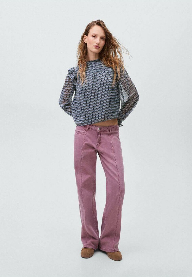 Blusa a trama blu e bianca con volant sulle spalle, abbinata a pantaloni rosa a vita alta. La modella si trova su uno sfondo chiaro.