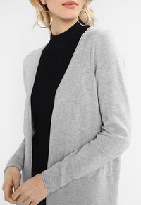 Svetle šedý cardigan vyrobený z měkké pleteniny, otevřený vpředu, s dlouhými rukávy a žíhanou strukturou na lemu a rukávech.