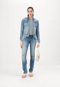 Veste en jean bleu clair, chemise rayée, jeans bleu clair, sandales beiges, portant un petit sac à main blanc. Tenue décontractée au design simple.