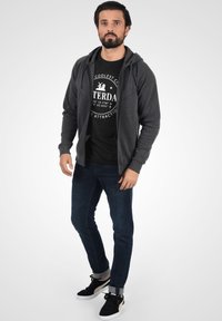 Grå zip-up hættejakke over en sort t-shirt med hvid grafik, parret med mørkeblå jeans og sorte sneakers med hvide accenter.