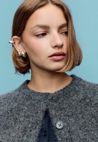 Siv pulover iz teksturiranega pletenega materiala z zaobljenim izrezom, na katerem sta dva črna gumba, nošen preko temno modre majice. Enostavna zasnova in mehak material.