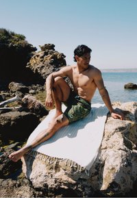 Osoba sjedi na stijenama pored vode, odjevena u šarene kupaćice s uzorcima. U pozadini se vidi bistra voda i kamenita obala.