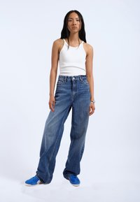 White ribbed tank top, high-waisted blue wide-leg jeans, and blue sneakers, set against a neutral background.