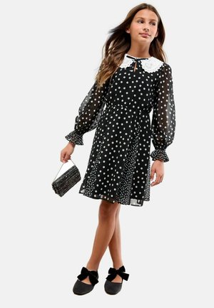 Jeune fille portant une robe noire à pois avec des manches transparentes, un col en dentelle blanche, des chaussures noires avec des nœuds et tenant un petit sac à main noir.