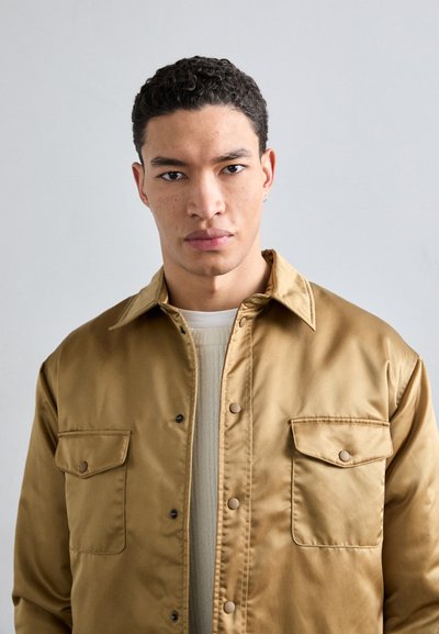 Brown satin jacket with a collar, two front pockets, and snap buttons, worn over a cream sweater, featuring smooth texture and a relaxed fit.