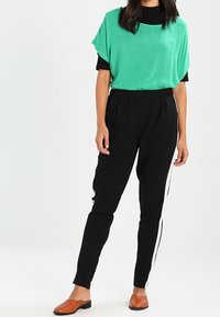Top verde a maniche corte sovrapposto a una maglietta nera a maniche lunghe, abbinato a pantaloni neri a taglio stretto con strisce bianche laterali e scarpe marroni slip-on.