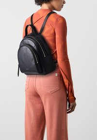 Svart skinnsekk med tre glidelåsrom, teksturert finish, slanke stropper og gullfarget hardware, båret på ryggen.