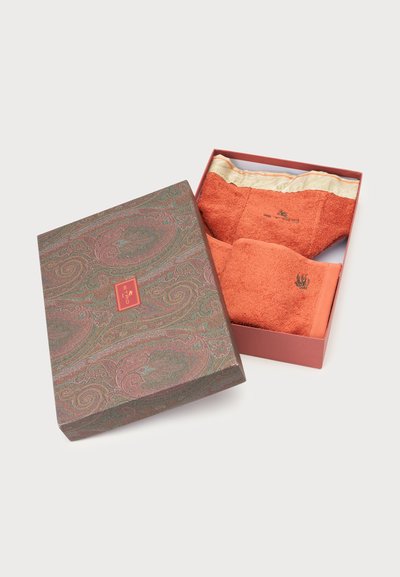 Boîte cadeau avec deux serviettes orange brûlé pliées, dont l'une avec une bordure dorée, placées dans un couvercle à motif paisley posé à côté de la boîte.