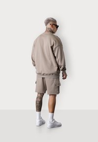 Sweatshirt beige surdimensionné et shorts cargo assortis en tissu doux. Baskets blanches et tatouage décoratif visible sur la jambe.