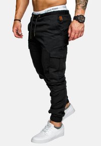 Schwarze Cargo-Jogginghosen mit einem Kordelzugbund, seitlichen Taschen und tapered Knöcheln. Kombiniert mit weißen Sneakers und einer Uhr.