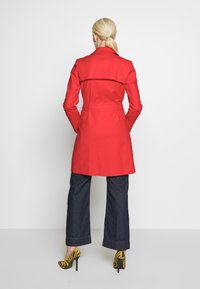 Trench-coat rouge avec une taille cintrée par une ceinture et un détail d'overli au dos, porté sur un jean denim foncé à jambes larges et des escarpins à talons hauts rayés jaunes et noirs.