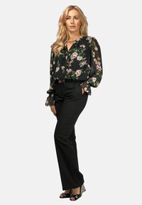 Czarna bluzka w kwiaty z przezroczystymi rękawami, zestawiona z dopasowanymi czarnymi spodniami. Modelka ma na sobie buty typu peep-toe, które eksponują elegancki detal w postaci paska.