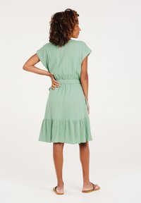 Hellgrünes, knielanges Kleid mit kurzen Ärmeln, Rüschensaum und einem taillierten Gürtel. Aus strukturiertem Stoff gefertigt. Keine sichtbaren Muster.