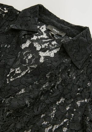 Schwarze Spitzenbluse mit Kragen, die ein florales Muster und eine durchsichtige Textur aufweist. Das Etikett lautet "NEXT." Der Stoff hat aufwendige Details.