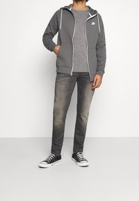 Sudadera con capucha gris con cierre y detalles en blanco, camiseta gris y vaqueros grises descoloridos. Zapatillas negras completan el conjunto. Fondo liso.