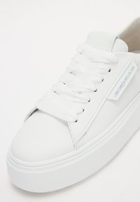 Sneakers en cuir blanc avec des lacets plats blancs, un bout arrondi et une semelle épaisse blanche. Présente un petit détail de logo bleu sur le côté.