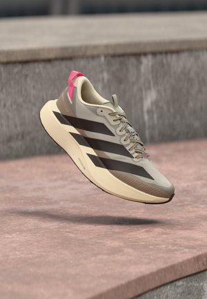 Chaussure de course beige clair avec des rayures noires, une semelle beige et un onglet de talon rose flottant au-dessus d'une surface en béton rose.